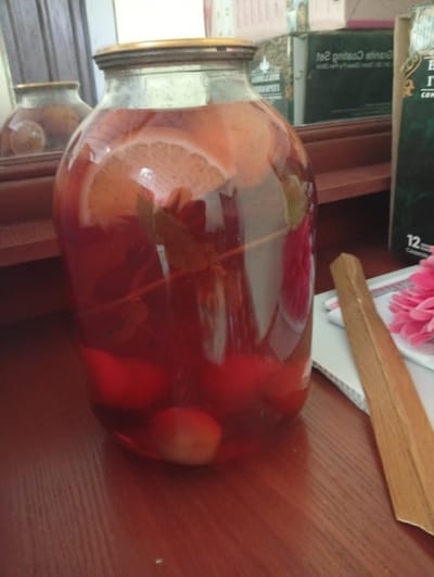 A large glass jar filled with a red liquid and pieces of fruit, possibly oranges and berries, sits on a wooden surface. The jar is topped with a golden lid. In the background, a mirror reflects another similar jar. Further back, there are some books or boxes with text on them. One book, in particular, has a green cover and the number "12" is clearly visible on it. To the right of the jar, a wooden stick rests on a white surface, next to a bright pink flower, possibly a dahlia. The overall lighting suggests it could be daytime. The scene appears to be indoors, likely in a home setting, possibly in Akaltyn, Uzbekistan.