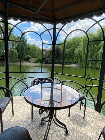 A glass-topped table with a metal base sits on a patterned tile patio. The table is reflecting the sky, which is filled with white, fluffy clouds. Behind the table, a body of green water is visible, likely a pond or lake, surrounded by lush green trees. The scene appears to be set in an outdoor gazebos or covered patio area, with a decorative metal railing. The time of day appears to be daytime, with bright sunlight casting shadows. The weather seems pleasant, with a clear sky and evident clouds. There are no people or text visible in the image. The overall impression is one of a peaceful, natural setting, possibly a restaurant or cafe with an outdoor seating area overlooking a scenic view.
