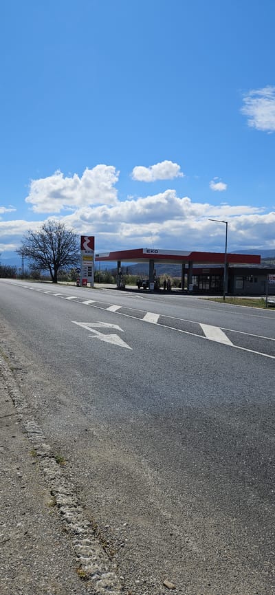 A slightly rotated shot of an EKO gas station along a road in Usoika, Bulgaria. The gas station has a red roof and an adjacent building. The road has directional arrows painted on the surface. A bare tree stands near the gas station's price sign. The sky is blue with scattered cumulus clouds. The foreground is dominated by the asphalt road surface.