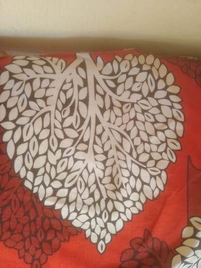 The image shows a close-up view of a red patterned fabric, likely a pillowcase or bedsheet, featuring a stylized tree with heart-shaped leaves. The tree's branches and some leaves are depicted in white and brown, creating a striking contrast against the red background. The fabric appears to be lying on a surface, with a wall visible in the upper portion of the image. The location is identified as Majemu, Nigeria, but no specific urban context is discernible from the image itself.