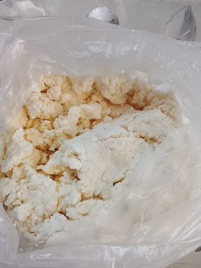 This is a close-up, overhead shot of a large quantity of raw, unprocessed cottage cheese, possibly being stored or transported in a clear plastic bag. The cottage cheese has a clumpy, uneven texture, with some pieces appearing slightly drier and lighter in color than others, suggesting varying moisture content. The plastic bag is translucent, allowing the texture and color of the cottage cheese to be clearly visible. The background is out of focus, but appears to be a light-colored, possibly metallic surface, suggesting a kitchen or storage environment. The image does not provide any information about the city of Kerben, Kyrgyzstan, or any specific details about people, actions, or interactions.