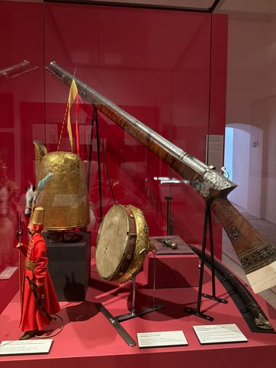The image displays a museum exhibit featuring historical artifacts within a glass case against a red background, likely at the Bavarian Army Museum in Ingolstadt, Germany. The main subjects are an ornate long gun, a decorative hand drum (Handpauke), a gilded helmet or hat, and a historical figurine.
The long gun, prominently displayed, features an elaborately decorated wooden stock with metal inlays and a detailed lock mechanism. Below it, a gilded helmet or hat, intricately patterned, is presented with feather-like adornments. To the right, a round hand drum with a skin head and studded decorations, identified by its label as a "Handpauke, um 1600/20," is visible. It is described as being made of "Kupfer, vergoldet, Türkise, Rubine" (copper, gilded, turquoise, rubies) and donated by the "Verein der Freunde des Armeemuseums, München, 1935." Below the drum, a figurine depicting a historical figure, possibly an Ottoman infantryman, is dressed in a red coat with gold trim, holding a sword.
The artifacts are held in place by black metal stands. Visible information labels in German provide details about the objects. One label for the hand drum specifies its approximate date as "around 1600/20," its materials, and its origin from the "Bayerisches Armeemuseum" (Bavarian Army Museum) with inventory number "A 10841." Another partially visible label to the right of the gun mentions "17. Jahrhundert" (17th Century) and "München, 1864." The scene is a static museum display, presenting historical items for public viewing. Reflections on the glass case indicate an indoor setting.