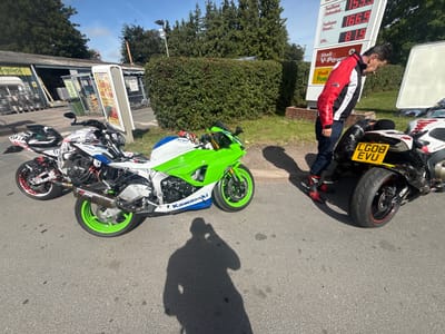 At a petrol station in Much Dewchurch, United Kingdom, two sports motorcycles are parked side-by-side on an asphalt surface. The motorcycle on the left is predominantly white with red and black graphics and has a green rim on its front wheel. The motorcycle on the right is black and yellow with a prominent license plate reading "LG08 EVU". A man wearing a red, black, and white jacket, blue jeans, and black and red motorcycle boots is bending over the motorcycle on the right, appearing to be inspecting it. Behind the motorcycles is a hedge, and further back, a building with a sign reading "TopSpec" is visible, suggesting this is a commercial area or a shop. A petrol price display board is also present, showing prices for Unleaded, Diesel, and LPG. The sky is clear and blue, indicating a sunny day.