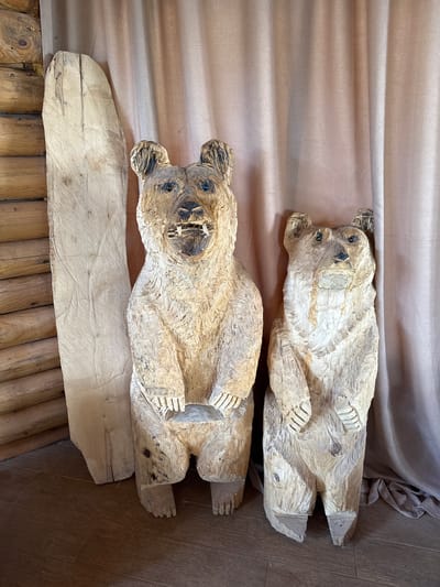 Two carved wooden bear figures lie horizontally on a surface. The bottom bear rests on a wooden plank, which is on a wooden floor. The top bear rests on a light tan cloth. The scene is located in Gornje Mrke, Montenegro.