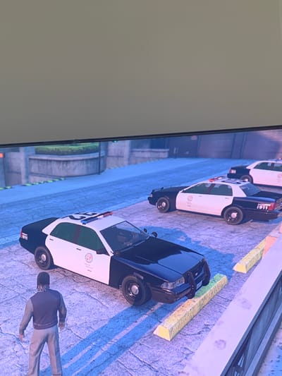The image shows a scene from a video game, likely Grand Theft Auto V, given the police car models and graphics. The scene depicts a row of police cars parked on a street. The cars are black and white and have "1911" written on their rear fenders. The ground is paved with a textured surface, and there are yellow concrete barriers alongside the road. In the foreground, a person is lying on the ground, wearing a dark jacket and light-colored pants. The person appears to be male and has braided hair. The overall atmosphere suggests a scene of law enforcement activity or a potential crime scene within the game. The lighting suggests it could be daytime. There is no discernible information about weather conditions or specific emotions. The location context of Vejprty, Czechia, is not directly evident from the visual elements of the image.
