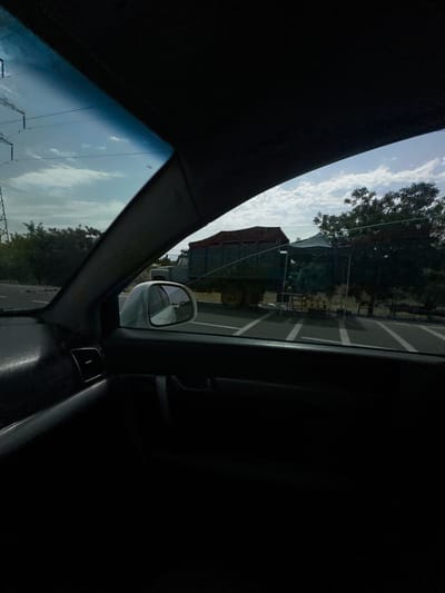The image is taken from inside a car, looking out the passenger side window. The car's interior is dark and partially visible, with a black dashboard and a silver vent. The side mirror is visible, reflecting the sky. Outside the window, a building with a red roof is visible, as well as trees and a road. The sky is partly cloudy, and the sun appears to be setting, casting a warm light. There are power lines visible in the distance. The scene appears to be a rural or suburban area, possibly in Istiqlol, Uzbekistan, given the location context. No people are visible in the image.