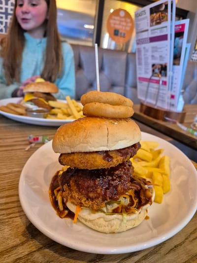The image shows a plate of food with a burger, fries, and onion rings. The burger appears to have a double patty, and there is a skewer holding the onion rings in place. The food is on a white plate, which sits on a wooden table. In the background, a girl is visible, holding another plate with a similar meal. There is also a menu stand and other dining-related items in the background. The setting seems to be a restaurant in Breckland District, United Kingdom.