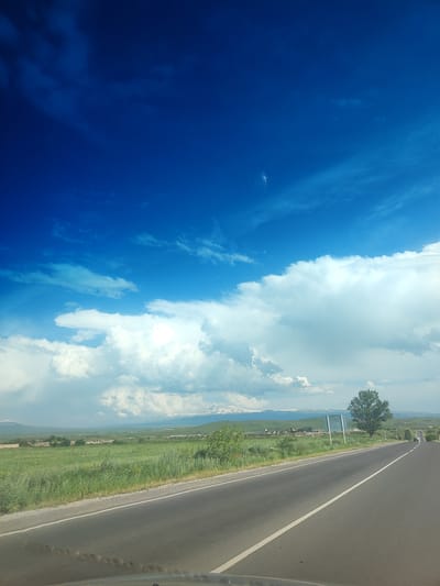 The image captures a scenic view of a road journey through a rural landscape in Azatan, Armenia. The scene is dominated by a vast, vibrant blue sky filled with dramatic white cumulus clouds, suggesting fair weather and possibly a warm, sunny day. Below the expansive sky, a long, winding asphalt road cuts through a lush green field. The field is dotted with vegetation, and in the distance, there are signs of human habitation, including scattered buildings and what appear to be farm structures.
Further in the background, a range of mountains is visible, with some peaks possibly capped with snow, indicating a significant elevation or a cooler climate in the distance. A solitary, mature tree stands prominently to the right of the road, adding a touch of natural beauty to the composition. A few vehicles are seen on the road ahead, suggesting active travel. The overall mood is one of tranquility and the freedom associated with a road trip through open country. The perspective is from within a vehicle, looking forward and slightly upwards, implying the photo was taken while driving. There is no visible text in the image.
