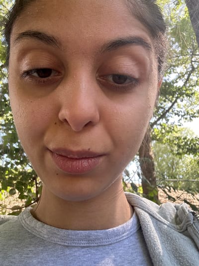 A close-up, downward-facing selfie shows a person's face and upper chest against a blurred background of green foliage and tree trunks, suggestive of an outdoor, wooded environment, possibly a park or garden. The individual, appearing to be female with dark hair pulled back, has her eyes partially closed and a neutral expression. Her skin shows a slight sheen, indicating perspiration or humidity. She is wearing a grey t-shirt and a grey hooded sweatshirt. The image does not contain any discernible elements that would place it specifically within the city of Augsburg, Germany.