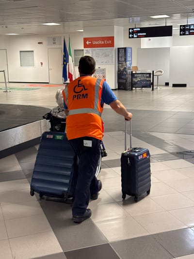 The image depicts an airport scene, presumably in Cinisi, Italy, based on the signage. A man wearing a high-visibility orange vest with "PRM" and a wheelchair symbol stands near a baggage carousel. He is pulling two blue suitcases. Behind him are the Italian and European Union flags. Signage in the area includes "Uscita Exit," "Dogana Customs," and advertisements for "Sicilia Outlet Village."