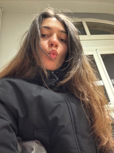 A young woman with brown hair puckers her lips for the camera. She wears a black jacket, and her hair is tousled across her face and shoulders. A window with white trim and an arched top is visible in the background. The overall lighting suggests an indoor setting, likely at night. The photo is taken from a slightly low angle and is slightly rotated.