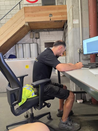 A man is seated at a desk, writing on a piece of paper. He is wearing a black polo shirt and dark shorts. A yellow and green high-visibility vest is draped over the back of his black office chair. The man is working in what appears to be a warehouse or industrial setting. In the background, there is a large computer monitor displaying a spreadsheet, a keyboard, and a telephone. There are also shelves with storage bins, a wooden staircase, and a large red pipe. The image was taken in Zeppelinheim, Germany.