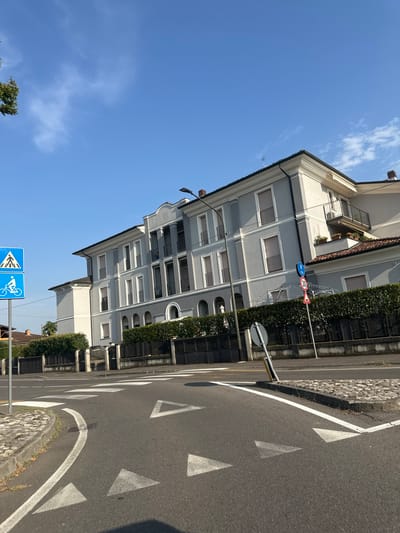 The image shows a street scene in Rudiano, Italy, on a clear day with a blue sky and scattered white clouds. In the foreground, the asphalt road curves to the left, marked with white directional arrows and a pedestrian crossing. A paved curb with cobblestones lines the edge of the road.
To the left of the road, a blue sign with a white pedestrian symbol above a white bicycle symbol is mounted on a pole. Further left, a tree with green foliage is partially visible.
Dominating the background is a large, multi-story building with a light gray facade, white trim, and dark balconies. The building has multiple windows with closed shutters. A manicured hedge forms a barrier in front of the building, behind which a dark metal fence is visible.
On the right side of the road, another pole displays two traffic signs: a circular sign with a red border and the number "30" in black, indicating a speed limit, and a triangular sign with a red border and a black symbol that appears to be an exclamation mark, warning of a hazard. A lamppost is also visible near the building. The overall scene depicts a residential or commercial area in a town.