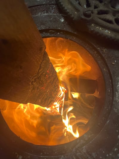 A close-up image shows a log burning inside a wood-burning stove. The stove is made of dark metal, with decorative ironwork visible on the left. Bright orange and yellow flames engulf the log, which is positioned diagonally across the opening. The interior of the stove is a warm, glowing orange color. The flames dance and swirl, creating a dynamic and intense heat. The wood itself has a charred, textured surface.