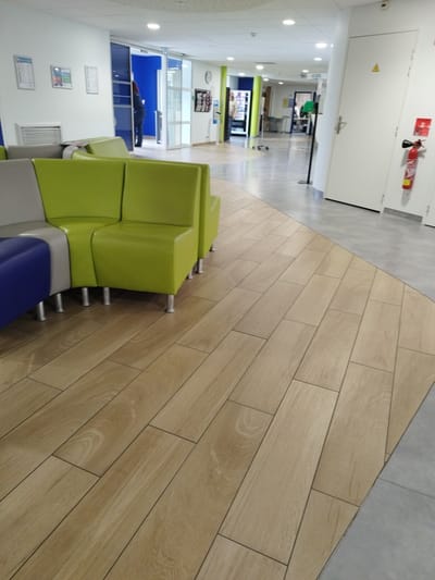The media file captures an interior shot of a modern, well-lit hallway or waiting area, likely in a public building such as a hospital or office complex, given the presence of a fire extinguisher and a sign that appears to be in French. The scene is dominated by a contemporary waiting area with modular seating in shades of blue, grey, and bright green. The flooring is a prominent feature, with a transition from a light-colored wood-effect laminate to a smooth grey surface.
In the background, a person is visible walking through a set of glass automatic doors into an adjacent brightly lit blue room, suggesting a movement or transition within the facility. Further down the hallway, other doors and what appear to be vending machines or information kiosks can be seen. The overall atmosphere is clean and functional.
Notable details include the bright, artificial lighting, indicative of an indoor environment during daytime. The fire extinguisher mounted on the wall, a safety feature, provides a clue to the setting's nature. The signage is partially obscured, but a yellow triangular warning symbol is visible on a white door. The location context, Cherbourg-en-Cotentin, France, suggests this is a public space within that region. No specific activity or event is clearly depicted, but the scene implies general use of the space as a passage or waiting area.