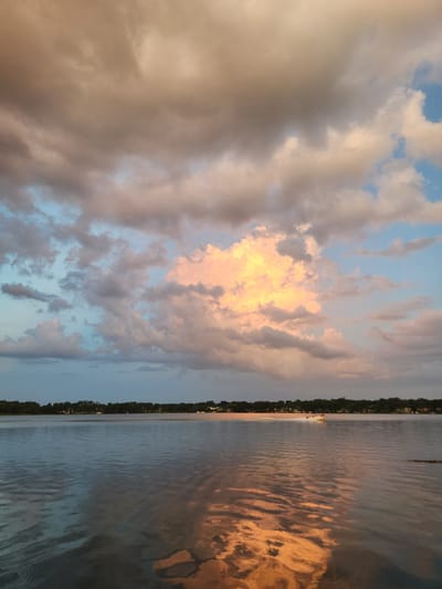 The image depicts a serene lake scene in Winter Park, United States of America, under a dramatic sky filled with colorful clouds. The sky is a canvas of soft blues, pinks, and oranges, with a prominent large cloud illuminated by sunlight, creating a warm glow. The water's surface reflects the sky's hues, adding to the tranquil ambiance. Along the horizon, a line of trees and scattered houses suggest a suburban or natural setting. A small white boat with people aboard is visible on the right side of the lake, leaving a wake as it moves. The overall impression is one of a peaceful evening at dusk.