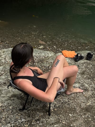 A woman with dark hair pulled into a bun sits in a portable chair by the edge of a body of dark, reflective water. She wears a black swimsuit and has a tattoo on her right thigh. Her arm is resting on her lap, and her bare feet are on the rocky ground. To her right are a folded orange shirt and a pair of black water shoes. The ground is covered in small, grey and brown stones. The water appears to be a river or lake. There is no specific indication of the location being in Khleborob, Russia.