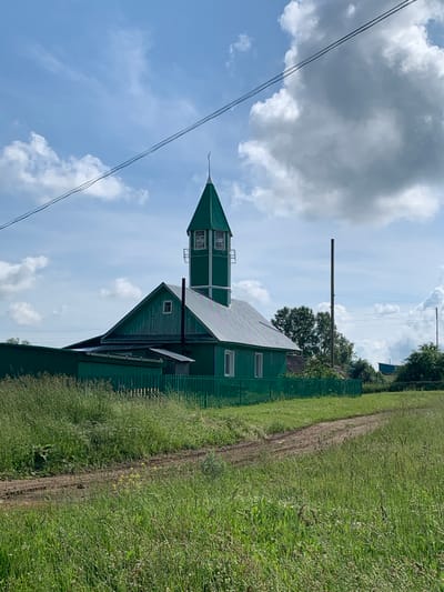 The image shows a bright green wooden building with a tall, narrow tower topped with a crescent moon and star, resembling a minaret. The building has a sloped roof and several windows. It is surrounded by a green wooden fence and tall grass. A dirt path leads through the grassy area in front of the building. The sky is blue with scattered white clouds. The building is located in the village of Barsai, Russia.
