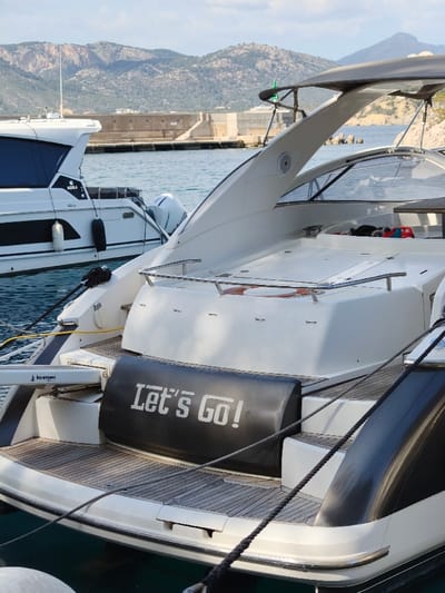 The image depicts two motor yachts docked in a marina in Santa Ponça, Spain, under a clear to partly cloudy sky. In the foreground, the stern of a white and dark grey yacht is visible, featuring a teak-style swim platform. A black inflatable fender or cover with "Let's Go!" in white lettering is positioned at the rear. Mooring lines, one yellow power cable, and a "Berenzoni" branded hydraulic arm are affixed to this yacht. Stainless steel handrails and a visible orange and white lifebuoy are present on the upper stern. Further back on this yacht, a clear or tinted windshield structure is evident. To the left in the mid-ground, a second white yacht, labeled "AQUILA" and powered by "MERCURY" outboard engines, is also docked, displaying black fenders along its hull. The calm, light blue-green water of the marina separates the vessels. In the background, rugged, treeless mountains rise, and a concrete breakwater or pier structure is visible across the water. No individuals are present in the scene.