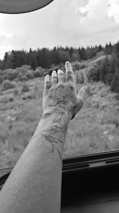 A black and white, close-up shot focuses on a tattooed forearm and hand extending from what appears to be a vehicle window. The tattoo, drawn in a delicate, vine-like pattern, adorns the arm and wraps around the wrist and onto the back of the hand. The fingers are slightly spread, reaching outwards. The background is blurred, showing a landscape of trees and foliage, suggesting an outdoor setting, possibly rural or natural, with a cloudy sky overhead. The dark frame of the vehicle window is visible in the foreground. The image does not provide specific details to pinpoint the location within Novaya Poyma (Nizhneyngarsky District), Russia.
