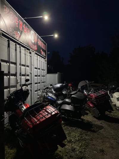 The image depicts a nighttime scene in what appears to be Doma 45 Kilometr, Russia, based on the Cyrillic lettering on a banner visible in the shot. Parked beside a large metal storage container are several motorcycles, mostly dark-colored touring models with red detailing and luggage racks. Two light poles illuminate the storage container, which features a banner displaying the words "ROARISH SOTREK." The surrounding environment consists of dark, likely grassy ground. The background shows a dark, possibly wooded area beneath a dark blue sky.