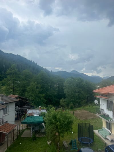 The image displays an elevated view of a rural residential area in Elhovets, Bulgaria, framed by forested mountains under an overcast sky. The upper portion of the frame is dominated by dense, grey cloud formations.

In the left foreground, a white two-story house with a brown tiled roof features a prominent chimney. Adjacent to it, a dilapidated wooden structure with a partially collapsed or unfinished roof is visible. Below these structures, a green tarp supported by poles creates a covered outdoor area with a concrete base, under which outdoor furniture including tables and chairs is arranged. A small, conical evergreen plant grows nearby, and a discarded white can rests on the surrounding grass.

The right foreground features a two-story house with a yellow facade and a red tiled roof. A balcony with a metal railing extends from its upper floor, holding several potted plants. A white satellite dish is mounted on the wall below the balcony. On a paved area adjacent to the house, a light green outdoor sofa and a table covered with a white tablecloth provide additional seating. A blue trampoline enclosed by a black safety net stands on a manicured grassy lawn, alongside a small blue and green plastic slide. A white bird is visible on the grass to the left of the trampoline. Part of a dark-colored vehicle is discernable at the bottom right, behind a wire fence. Beyond the lawn, a rectangular plot of tilled earth with visible furrows indicates a cultivated garden area.

The midground is characterized by a thick expanse of various green trees, forming a natural buffer between the residential structures and the mountainous terrain. The background reveals multiple layers of mountains, densely covered in dark green trees, receding into a hazy distance under the cloudy conditions. No human figures are present or actively engaged within the scene.