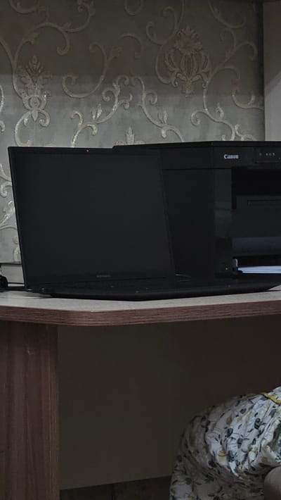 In Yandare, Russia, a black laptop is positioned on a wooden desk against a wall with ornate wallpaper. The laptop is open, displaying a dark screen, and appears to be powered on, indicated by a small red light visible on the bezel. To the right of the laptop, a black Canon printer is present. The environment suggests an indoor setting, possibly a home office or study. The lighting is dim, with the primary light source appearing to be ambient room light.