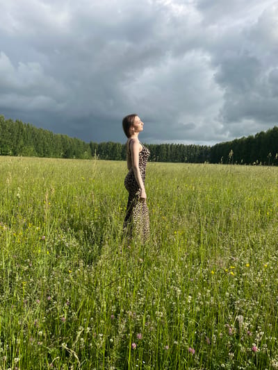 A young woman stands in a field of tall green grass and wildflowers under a cloudy sky. She is wearing a leopard-print dress and is facing towards the right side of the frame with her eyes closed and her face tilted upwards, seemingly basking in the sunlight. Behind her, a line of green trees stretches across the horizon, and the sky is filled with dark, dramatic clouds. The location is a meadow in Большой Гондыр, Russia.