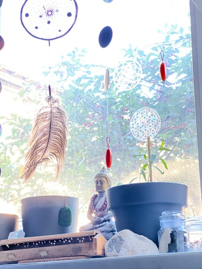 The image shows a windowsill with various decorative items. In the background, a window looks out onto a tree with red berries.

In the foreground, on the windowsill, there are several objects:
* A dark gray plant pot containing a small green plant.
* To the left of this pot, another dark gray pot is partially visible.
* A Buddha statue is positioned behind the pots, adorned with a beaded necklace.
* To the left of the Buddha statue, a green pendant is hanging from a string.
* A wooden incense holder with decorative cutouts and small elephant figures is in front of the pots.
* Several crystals, including a large white one, are arranged on the windowsill.
* A glass jar with a lid contains various black and white objects, possibly crystals or stones.

Hanging from the top of the window are several ornaments:
* A large dreamcatcher with a dark purple rim and intricate webbing.
* Several dark circular or oval shapes.
* A long, brown feather.
* Two small, red, oval-shaped pendants.
* A circular, geometric ornament that appears to be made of glass or acrylic, possibly a "Flower of Life" symbol.

The image does not provide any information about the city of Rezina, Moldova.