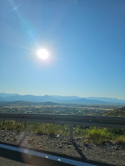 The image captures a panoramic view of a valley and distant mountains under a clear blue sky. The sun is prominently visible in the upper left quadrant, emitting lens flares. In the foreground, a silver guardrail separates the viewer from a rocky embankment covered with sparse vegetation and dry grass. The guardrail is supported by metal posts, one of which casts a shadow on the ground. Below the guardrail, a road surface with a white line is visible.

The valley in the mid-ground shows a sprawling settlement, consistent with the description of Gornji Kokoti, Montenegro. Buildings are scattered across the landscape, interspersed with patches of green and brown. The settlement extends towards the base of rolling hills and a more distant range of hazy, blue mountains that form the backdrop of the scene. The overall impression is one of a bright, sunny day with a vast landscape.