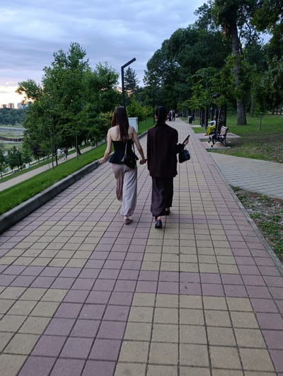 Two women walk away from the camera on a paved pathway in a park. The woman on the left wears a black strapless top and loose-fitting beige pants, carrying a black shoulder bag. The woman on the right wears a dark brown suit with wide-leg pants and carries a black handbag in her left hand, and a phone in her right. They are holding hands. The pathway is made of light yellow and pinkish-purple rectangular paving stones. To the left of the pathway, a grassy slope descends, with trees and buildings visible in the distance under a cloudy sky. To the right, the pathway continues alongside a grassy area with trees, park benches, and a lamppost. The overall scene suggests an evening stroll in a well-maintained urban park.