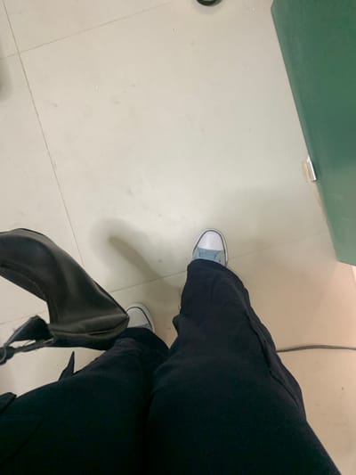 The image presents a top-down view of a person's lower body within an indoor setting. The individual is wearing black trousers and light-colored sneakers; the left shoe is white with a light blue upper, while the right shoe is primarily white. A black handbag rests on the light beige tiled floor next to the person's left leg. The floor shows visible grout lines and minor surface dirt. A dark green vertical surface, likely a wall or partition, is visible along the right side of the frame, featuring a small white fixture near its base. A thin dark cable or wire is present on the floor towards the bottom right. The specific location within Lauro De Freitas, Brazil, cannot be determined from the provided visual information.