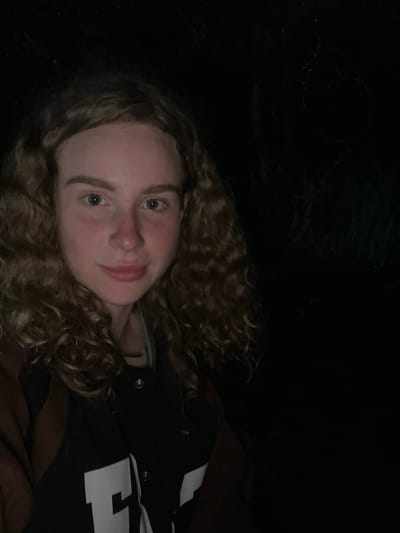 A young woman with curly hair is lying down, looking at the camera. She is wearing a dark jacket with a large white "H" on it. The background is dark and appears to be outdoors, possibly at night. The specific location is not discernible.