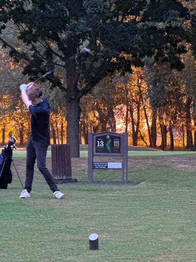 A man in black pants and a blue shirt is captured in the middle of a golf swing. He is on a golf course with a tee marker for the 13th hole nearby. A golf bag sits on the ground beside him. Trees with golden sunlight filtering through the branches are visible in the background, suggesting the photo was taken during sunset or sunrise. The lighting gives the image a warm tone.