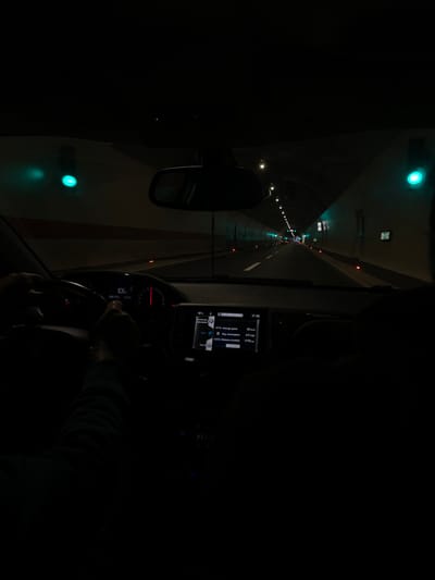 The scene depicts the interior of a vehicle moving through a tunnel in Mateševo, Montenegro. The perspective is from the front passenger compartment, looking forward through the windshield.

A driver's hands are positioned on a dark steering wheel. The instrument cluster shows "106" prominently, indicating speed, and "266.2km" for a trip distance. A central infotainment screen displays "18°C," the time "17:02," an average speed of "42 Km/h," an average fuel consumption of "4.7l/100," and a total calculated distance traveled of "1175 km." A dark rearview mirror is suspended from the ceiling. The indistinct, dark form of a second occupant is visible in the right foreground, consistent with a front passenger.

The tunnel interior features light-colored walls with a continuous red horizontal stripe at a lower level. The tunnel ceiling contains an array of recessed white lights extending into the distance. Large, circular green lights are affixed to both the left and right tunnel walls at regular intervals. The asphalt roadway comprises two lanes delineated by white markings. Small, orange-red reflective markers are present along the road's edges. The tunnel structure recedes into the background, maintaining consistent artificial illumination.