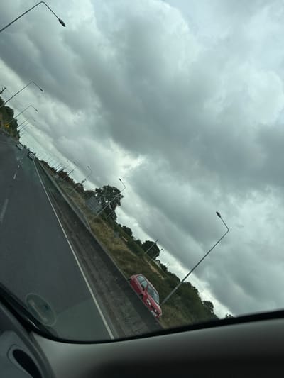 The image shows a view from a vehicle on a road with a cloudy sky overhead. In the distance, several streetlights are visible, lining the side of the road. There are trees and vegetation on either side of the road, with a red car parked on the right shoulder. The road appears to be a highway or major road with multiple lanes. The perspective suggests the image was taken while the vehicle was in motion. No people are clearly visible. The overall atmosphere is overcast, suggesting it may be raining or about to rain. The location is identified as being in the city of Slieverue, Ireland.