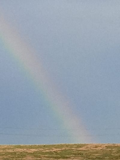 The image shows a rainbow arching across a light blue sky. The rainbow is positioned in the upper left quadrant of the frame and extends diagonally downwards towards the right. The sky is mostly clear, with a slightly overcast appearance. In the lower part of the image, a flat, grassy plain is visible. The terrain appears dry and sparsely vegetated. A faint, horizontal line, possibly a power line or a distant ridge, is present in the middle ground. The image does not contain any specific identifiable landmarks or features that would definitively place it in Yar-Bazar, Russia. There are no people or animals visible in the scene. The overall atmosphere is serene and natural.