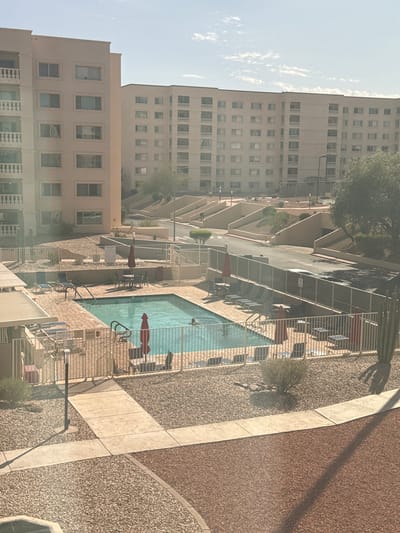 The image depicts an outdoor swimming pool area at a multi-story apartment complex in Scottsdale, Arizona, United States of America. The buildings are light-colored, with repeating balconies and numerous windows. In the foreground, a rectangular swimming pool with turquoise water is visible, surrounded by a light-colored paved deck. Several lounge chairs and patio umbrellas are placed around the pool area. A metal fence encloses the pool. A single person is in the pool. In the background, the apartment buildings dominate the scene, with terraced landscaping and a road. The sky is a pale blue with scattered clouds. The ground is covered with gravel and some desert vegetation, including a large saguaro cactus on the right side of the image. The lighting suggests it is daytime, likely mid-morning or late afternoon, given the shadows.