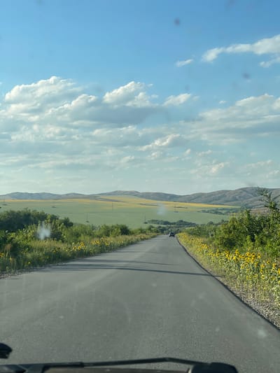 A view from a vehicle driving on a paved road towards rolling hills under a partly cloudy sky. The road is gray asphalt and lined with green vegetation and sunflowers on the right. In the distance, there are green and yellow fields with scattered utility poles. A dark-colored car is visible on the road ahead. The sky is blue with scattered white and gray clouds. The image appears to be taken in a rural area, possibly near the city of Progress, Kazakhstan, given the landscape.