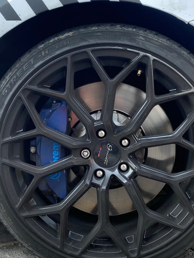 The image shows a close-up of a car wheel with a black Gianelle rim. The tire is a Street-H Hudi model, made in Vietnam. Behind the rim, a silver brake rotor is visible, and a blue Brembo brake caliper is seen through the wheel spokes. The image includes parts of the car’s body, one being white and another covered in black and white checkered pattern. The location of the image is not determined from the information given.