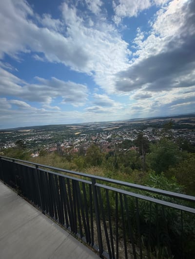 The image is a wide-angle, high-angle shot taken from an elevated position overlooking the city of Homberg (Efze), Germany. In the foreground, there is a concrete pathway and a dark metal railing. Beyond the railing, a dense canopy of green trees and foliage dominates the midground, obscuring much of the terrain immediately below.

Further into the distance, the cityscape of Homberg (Efze) is visible, characterized by numerous red-tiled roofs, white and cream-colored buildings, and the presence of a prominent church tower, likely St. John's Church, a well-known landmark in the city. The town extends across a valley, with houses nestled amongst trees.

The background features a vast, flat expanse of agricultural land, possibly fields of crops, stretching towards a hazy horizon. In the far distance, a few wind turbines are faintly visible, indicating the rural surroundings. The sky above is a dynamic mix of bright blue patches and scattered white and gray clouds, suggesting fair weather with some cloud cover. The overall impression is of a panoramic view of a moderately sized European town situated within a green, rural landscape under a partly cloudy sky.