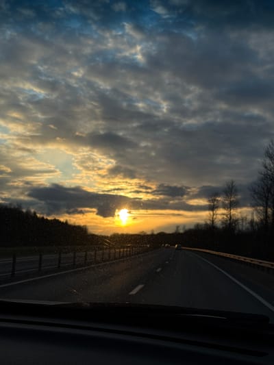The image shows a view from inside a vehicle, likely a car, traveling on a highway. The sky is filled with clouds illuminated by the setting sun, with the sun itself visible near the horizon. A tree line is visible between the sky and the highway. The highway has lane markings and a guardrail. The interior of the vehicle is visible on the right side of the frame. It is impossible to determine the location within Mshaga, Russia from the image alone.