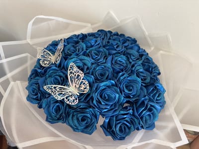 A bouquet of deep blue, glitter-covered artificial roses is arranged in a dome shape and wrapped in sheer white fabric. Two white, decorative butterflies with intricate cutouts are placed on top of the bouquet, one on the left and one towards the top right. The bouquet is set against a white background, likely a wall, with subtle shadows indicating ambient lighting. The image appears to be taken indoors, possibly in a residential setting. The location in Oława, Poland, is not discernible from the visual information provided in the image.