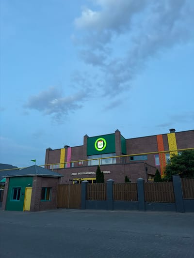 An exterior shot captures the Jolly International School, a brick building located in Orto-Sai, Kyrgyzstan. The school features a prominent green facade on the upper section of the building, adorned with a circular logo that reads "JIP". Vertical panels of vibrant colors, including yellow, pink, and orange, are integrated into the brickwork, suggesting climbing walls. Below the green facade, the text "JOLLY INTERNATIONAL SCHOOL" is visible. In the foreground, a small, green brick guardhouse with a yellow door and windows is situated to the left of a wooden fence. The sky above is a pale blue with scattered clouds.