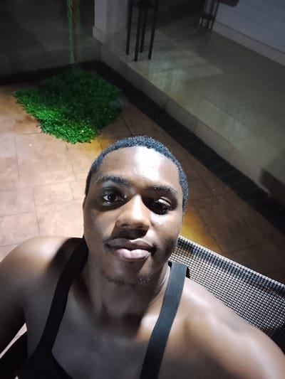 A male individual is depicted in a selfie, positioned in a dimly lit patio or interior space in Nibo, Nigeria. The subject is a dark-skinned male with short, dark hair, looking directly at the camera with a neutral expression. He is wearing a dark, sleeveless garment with thick shoulder straps visible on his shoulders. The upper right portion of a dark, textured object, possibly a chair back or railing, is visible behind his right shoulder. The flooring is composed of light brown tiles with dark grout lines. In the upper left background, a small, roughly square patch of artificial green foliage rests on the tiled floor. Further back, a light-colored wall or pillar is partially visible, with the dark legs of furniture (possibly chairs or a table) faintly discernible beyond it. Two thin, vertically hanging strands, one red and one green, are present in the far upper left background. Artificial light illuminates the scene, casting shadows on the subject's face.
