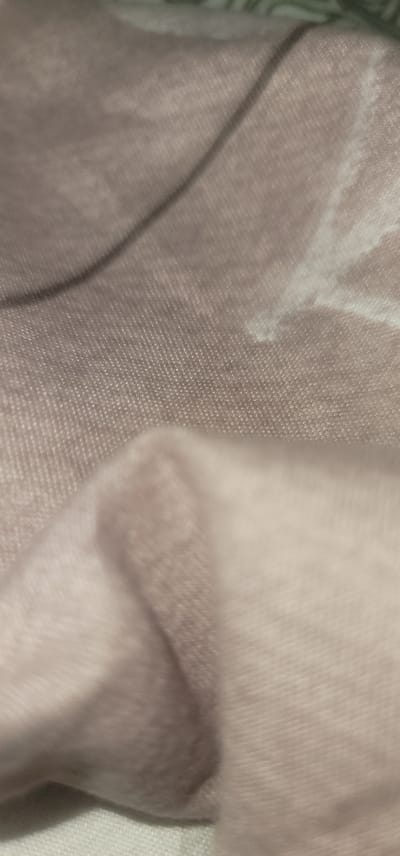 The image is a close-up, abstract shot of a textured fabric. The fabric appears to be a soft, light pink or mauve color and has a subtle weave pattern visible. There are folds and creases in the fabric, creating areas of light and shadow that give it a three-dimensional appearance. The lighting is soft, and the focus is shallow, blurring some areas of the fabric. There are no people, distinct objects, or identifiable environmental features visible in the image. The image does not depict any specific location in the city of Balykchi, Uzbekistan, or any other location.