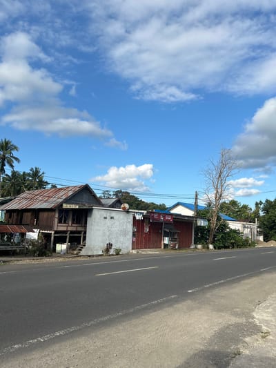 This is a vertical, eye-level shot of a roadside scene in Karama, Indonesia. The sky dominates the left side of the frame, a bright blue with scattered white, puffy clouds. A bare, dead tree stands starkly against the sky, its branches reaching upwards.
On the right side of the image, a paved road runs diagonally, with white and yellow lines marking the lanes. Along the side of the road are several buildings. The closest building has a rusty, corrugated metal roof and appears to be a small shop or dwelling. It has a red facade with some visible signage. Further back, a white building with a textured facade is visible, and behind that, more trees and structures can be seen.
The scene appears to be set during the day, with bright sunlight casting shadows. The weather seems fair, with clear blue skies and clouds. The presence of palm trees and the style of architecture suggest a tropical location like Indonesia. There are no people visible in the frame, and the overall atmosphere is one of quiet, rural life.
Visible text includes the word "KEMIL" on a red sign on one of the buildings.