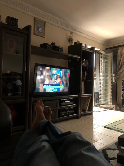 The scene portrays the interior of a residence in Soweto, South Africa, observed from a first-person perspective. The lower legs and bare feet of an individual, clad in blue jeans, are positioned in the foreground, suggesting a reclined posture. Directly ahead, a black Sony flat-screen television is centered on a dark brown entertainment unit. The television displays a live cricket match, featuring a batsman in a blue helmet and a scoreboard showing "BUTTLER SF" and a score of "124-3". Below the television, multiple audio/video components are stacked within the unit. Stereo speakers are placed on the upper shelves of the entertainment unit, with one on the far left and two centrally located above the television.
The walls are light-colored, featuring white decorative crown molding near the ceiling. A framed picture hangs on the wall to the upper left. To the right, a wooden door with an open, white metal security gate permits natural daylight to stream onto the light-colored tiled floor. A light brown curtain is visible beside the doorway. A small, dark decorative item is affixed to the wall near the ceiling on the right side. A portion of a glass-topped table is discernible in the lower right segment of the image.
