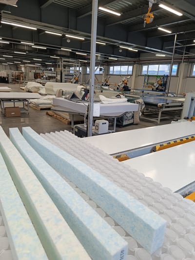 In an industrial facility, possibly in Zangiata, Uzbekistan, rows of foam mattress cores, some blue and white speckled, are arranged diagonally in the foreground. Behind them, a surface covered with white, individually pocketed springs stretches towards the background. Further back, in a large, well-lit factory floor with high ceilings and exposed ductwork, several large machines are visible. One machine appears to be a cutting or processing unit for mattress materials. A worker in a blue shirt is seated at one of these machines. Other stacks of white mattress components, fabric rolls, and industrial equipment are scattered throughout the space. The environment appears to be a manufacturing setting, likely for mattress production.