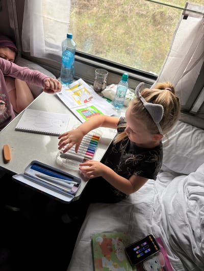 A young girl in a black flower-patterned shirt and gray cat-ear headband sits at a small table, drawing on a notebook. A woman in a pink sweater lies beneath the table. The table holds a box of markers, a pencil case with a ruler and pencil, and water bottles. Coloring books and a cell phone displaying a game are on the bed above. The background shows a window with blurred scenery. The location is not specified, and there is no evidence to suggest the image was taken in the city of Korovino, Russia.